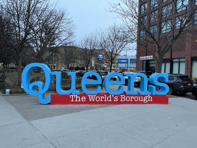 Large blue “Queens” sign with a red “The World’s Borough” base stands on a city sidewalk