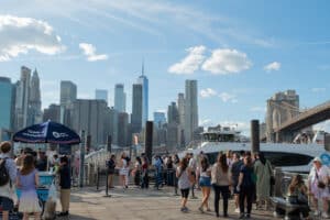 People Waiting to board the New York Ferry at the Brooklyn Bridge Ferry Landing. Visible in the background is the iconic One World Trade Center, along with the Brooklyn Bridge