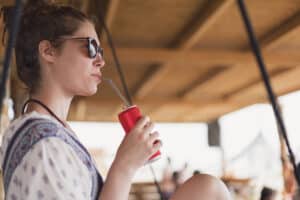 Attractive young woman relaxing on the swing at the beach bar and drinking soda.